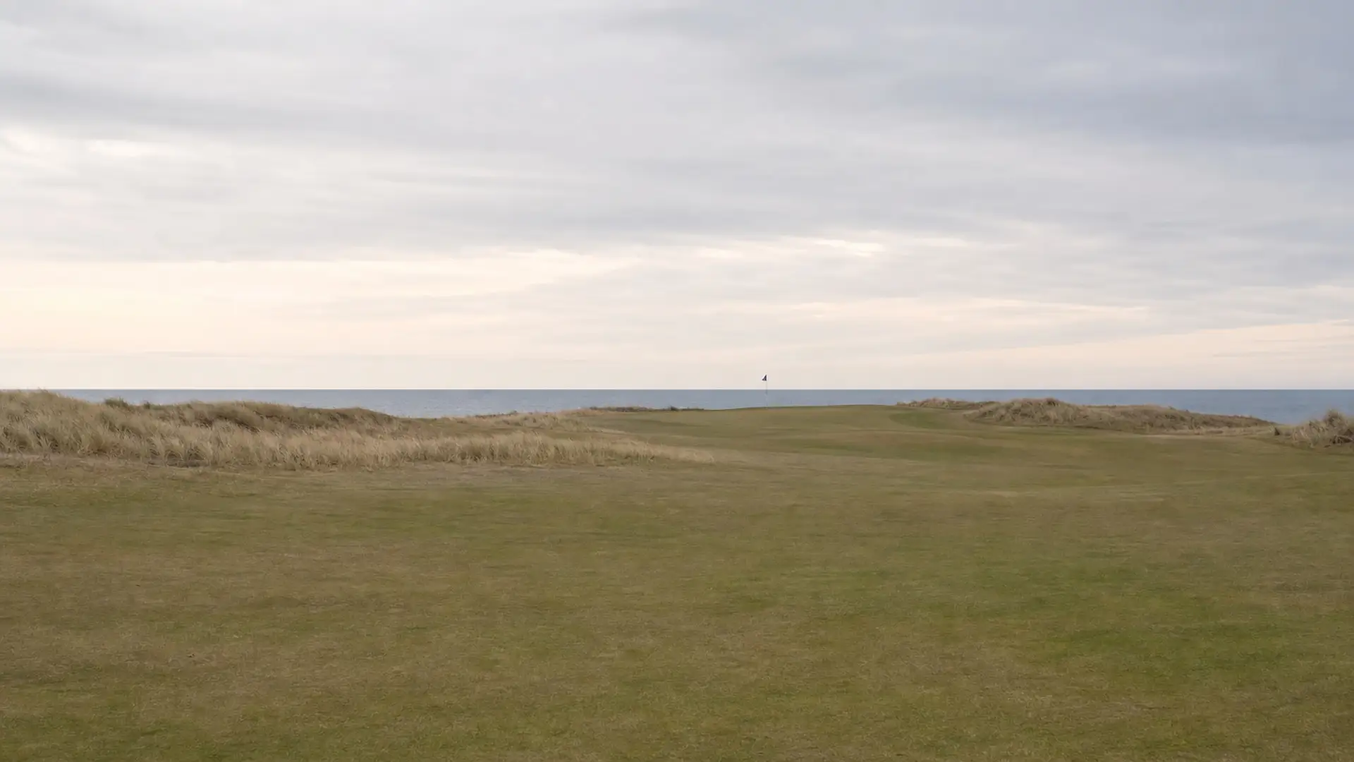 A flat coastal links course with marram grass in the rough and a flag against the sea horizon