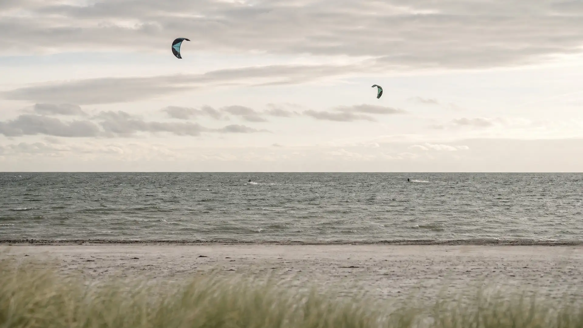 Two kites over the Öresund off a sandy beach, under a pale, broken sky
